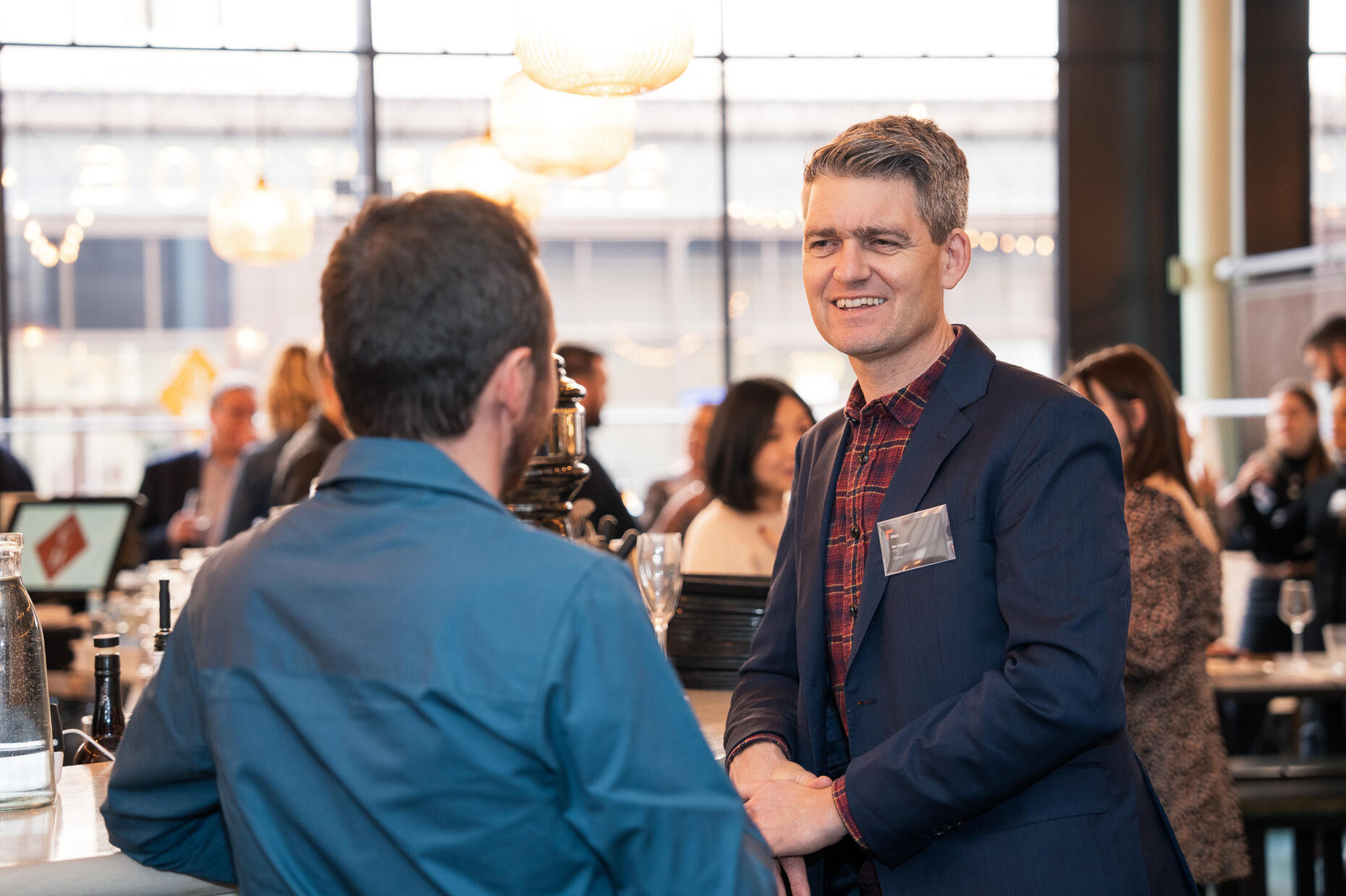 Ben Scoggins Man in a shirt and jacket standing and chatting to another man at a networking event